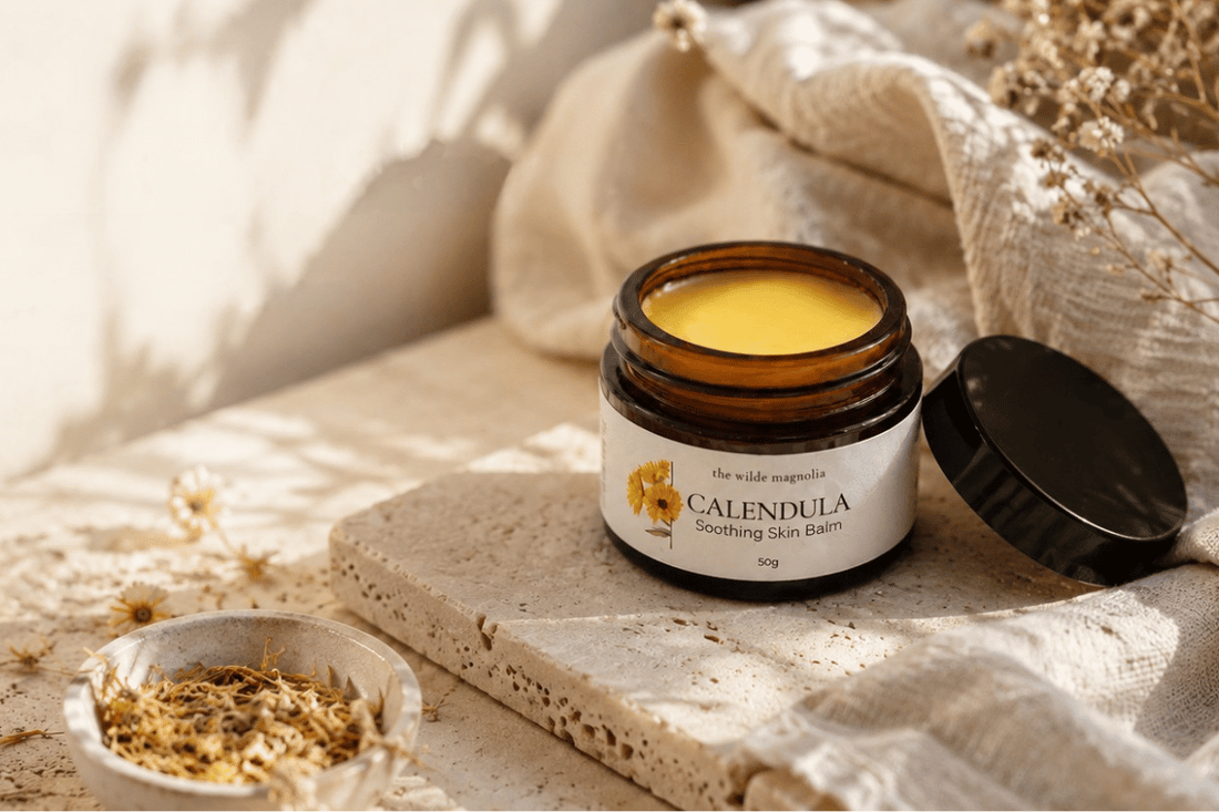 An amber jar of Calendula Balm by The Wilde Magnolia sitting on a travertine slab with dried flowers and a linen towel beside it