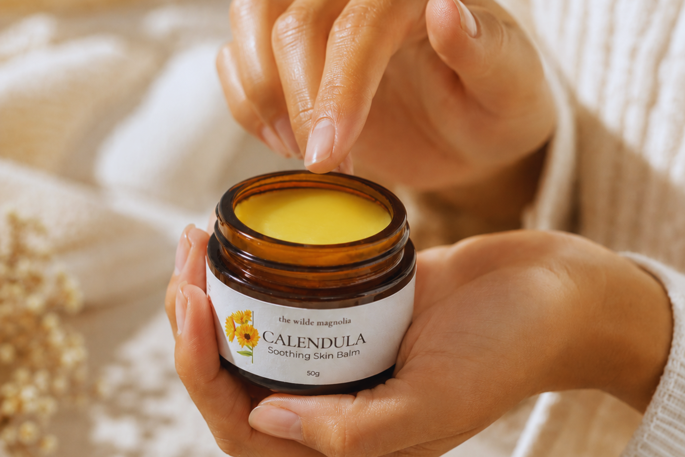 A jar of Calendula Soothing Skin Balm by The Wilde Magnolia held in a hand with fingers hovering over the top about to scoop some out