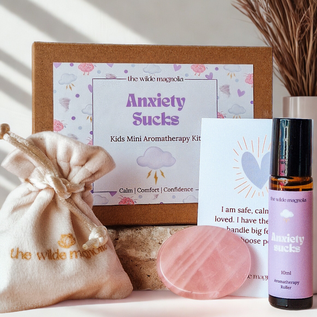 Kids mini aromatherapy kit with 'Anxiety Sucks' label and related items such as an amber glass anxiety sucks essential oil roller, affirmation card, rose quartz palm stone, and velvet pouch on a neutral background.