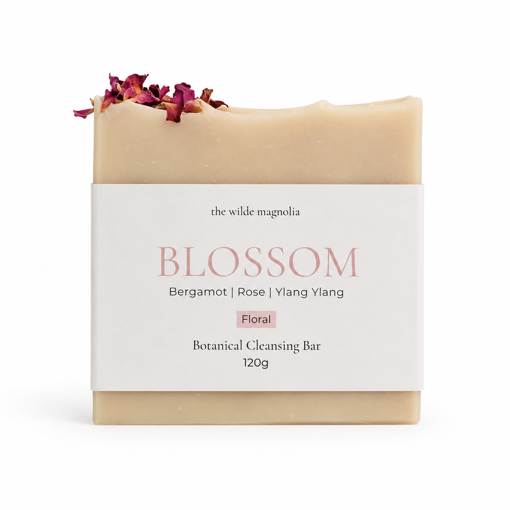 A bar of botanical cleansing soap from The Wilde Magnolia, named Blossom, wrapped in a white paper label with pink text. The soap bar itself is a warm cream-pink color and is decorated with dried rose petals on top.