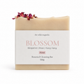 A bar of botanical cleansing soap from The Wilde Magnolia, named Blossom, wrapped in a white paper label with pink text. The soap bar itself is a warm cream-pink color and is decorated with dried rose petals on top.