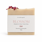 A bar of botanical cleansing soap from The Wilde Magnolia, named Blossom, wrapped in a white paper label with pink text. The soap bar itself is a warm cream-pink color and is decorated with dried rose petals on top.