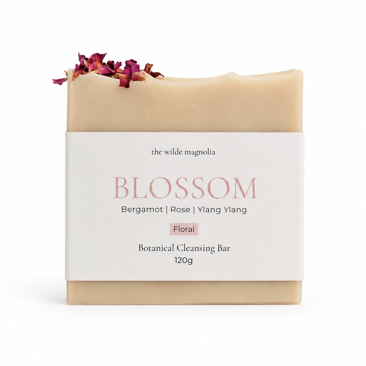 A bar of botanical cleansing soap from The Wilde Magnolia, named Blossom, wrapped in a white paper label with pink text. The soap bar itself is a warm cream-pink color and is decorated with dried rose petals on top.