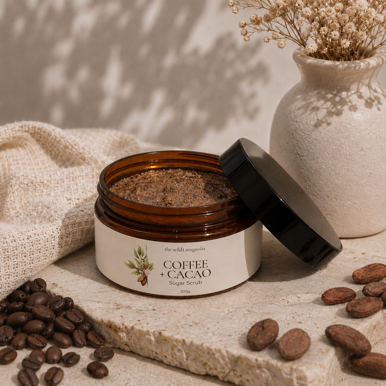 An open amber jar of Coffee + Cacao Sugar Scrub with its black lid leaning beside it, styled on a stone surface with coffee beans, cocoa pieces, a beige cloth, and dried flowers in a vase.