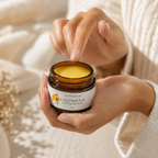 A person holding an open amber glass jar of Calendula Soothing Skin Balm, with their fingers hovering over the yellow balm inside. The background is a soft, warm beige with a cozy knit sweater visible on the right.