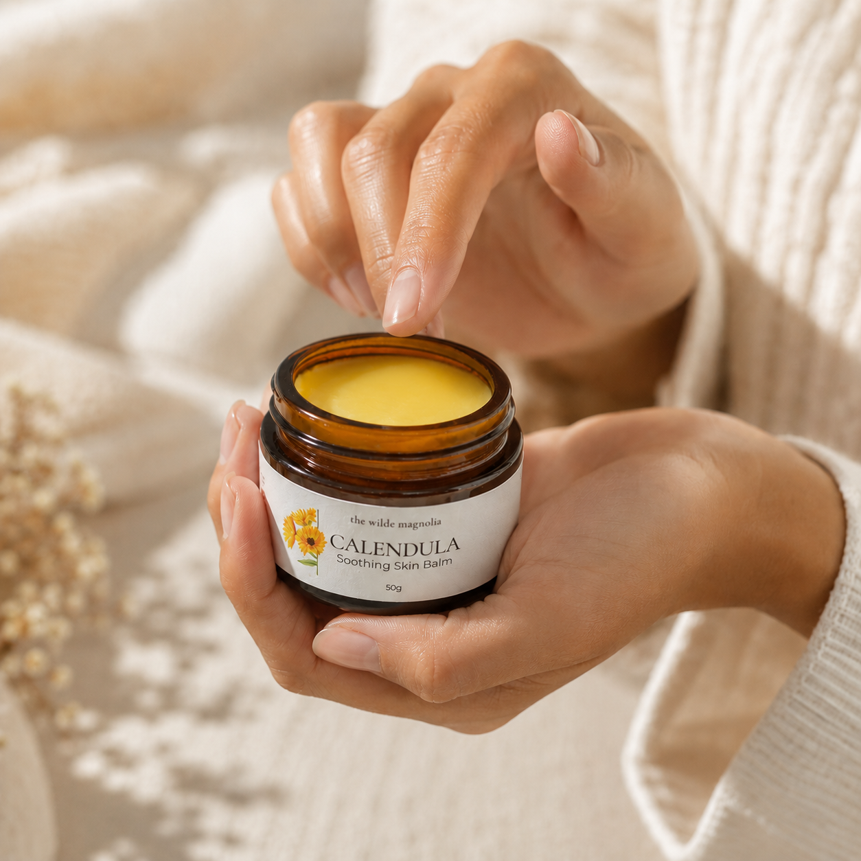 A person holding an open amber glass jar of Calendula Soothing Skin Balm, with their fingers hovering over the yellow balm inside. The background is a soft, warm beige with a cozy knit sweater visible on the right.