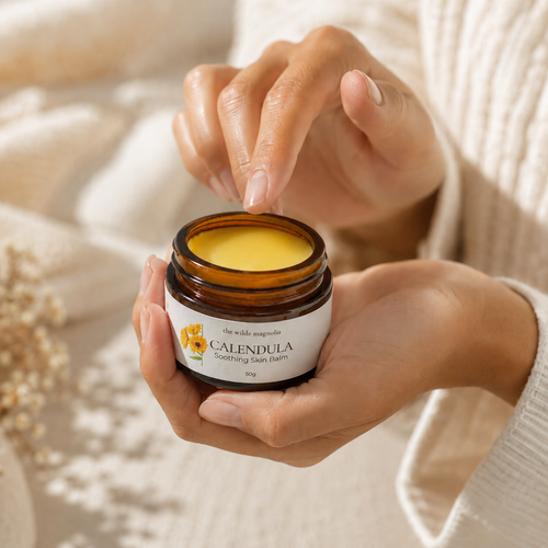 A person holding an open amber glass jar of Calendula Soothing Skin Balm, with their fingers hovering over the yellow balm inside. The background is a soft, warm beige with a cozy knit sweater visible on the right.