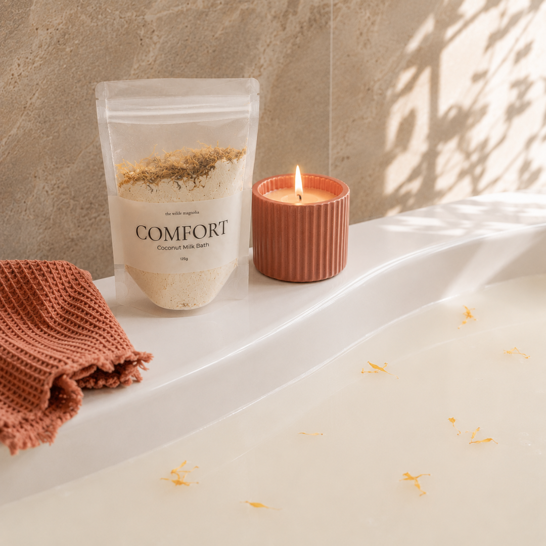 A clear stand-up pouch labeled COMFORT Coconut Milk Bath sits on a white bathtub beside a lit terracotta candle and a folded rust-colored towel, with warm sunlight and leaf shadows across the tub.
