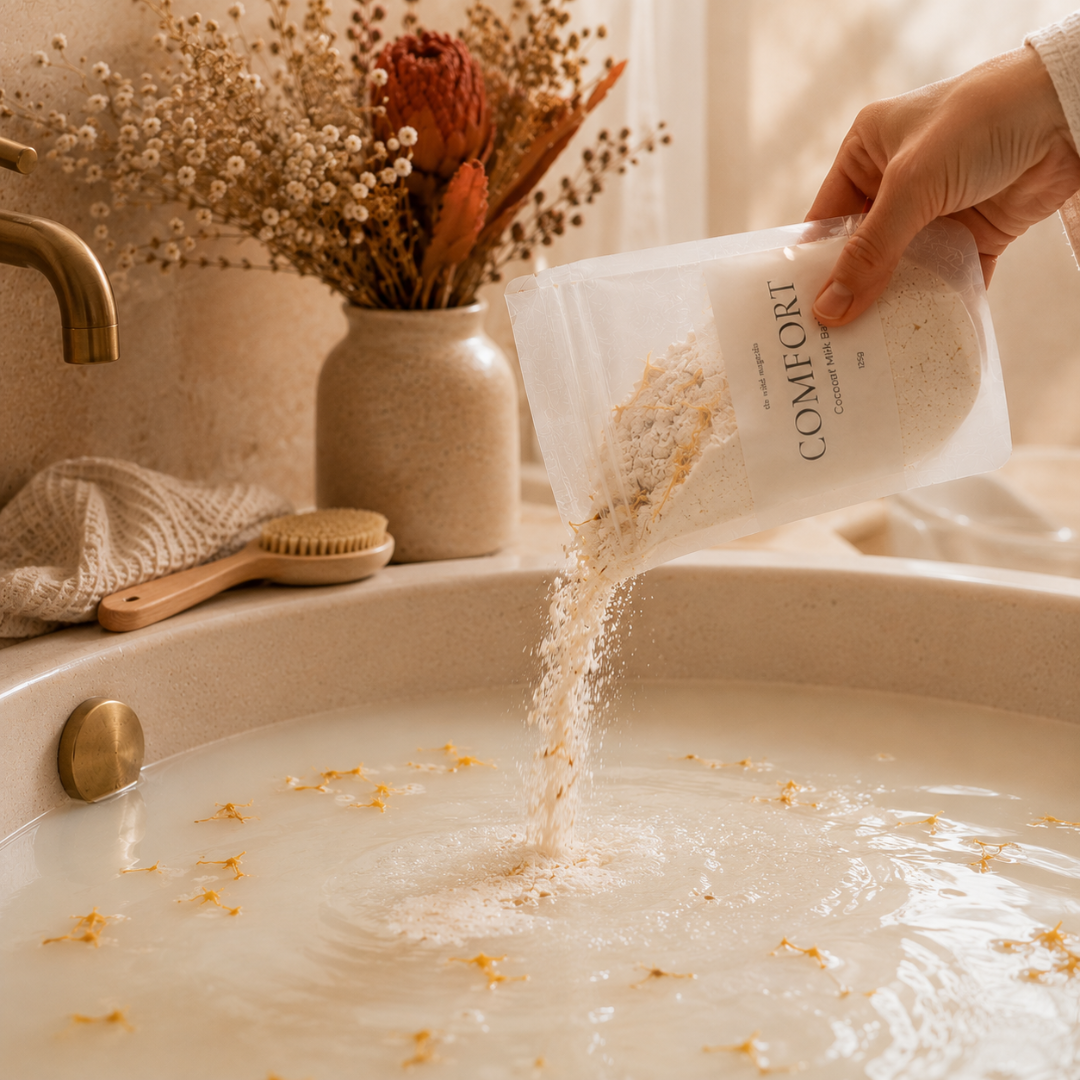 A hand holds a translucent pouch labeled COMFORT and pours a light beige powder into a warm bath. A rustic vase with dried flowers, a wooden brush, and a folded towel sit nearby in a softly lit bathroom setting.