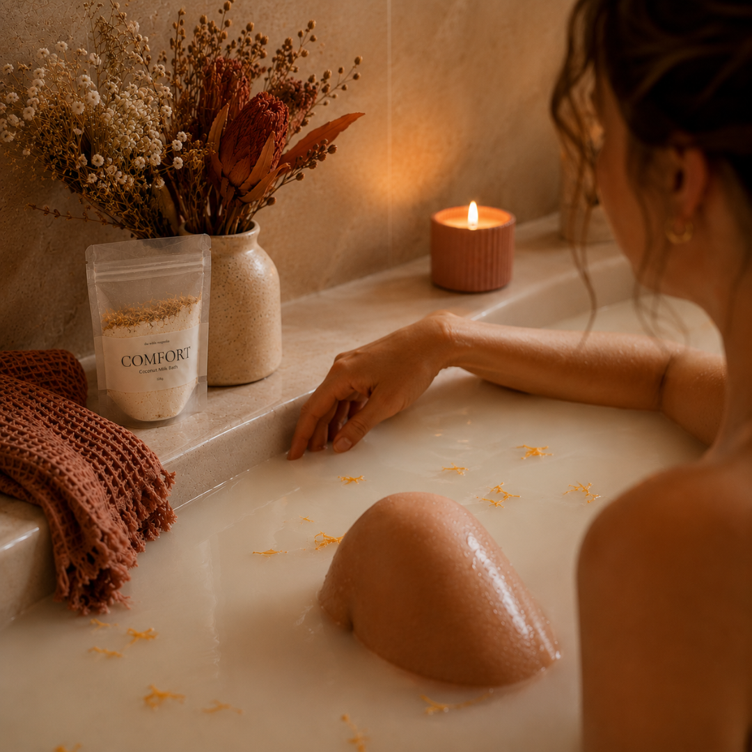 A person relaxes in a warm, dimly lit bathtub with a candle and dried flowers on the edge. A stand-up pouch labeled COMFORT sits on the ledge, suggesting a bath product in use.
