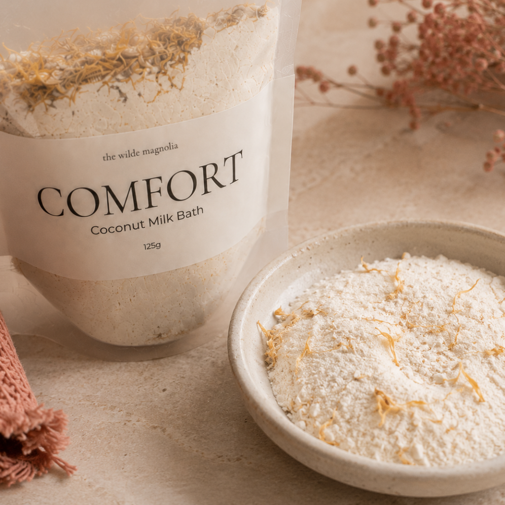A clear stand-up pouch labeled 'COMFORT Coconut Milk Bath' with a white paper label, shown beside a small ceramic bowl filled with the white bath powder and dried botanical pieces. The scene is styled on a beige surface with a folded pink knit fabric and dried flowers in the background.