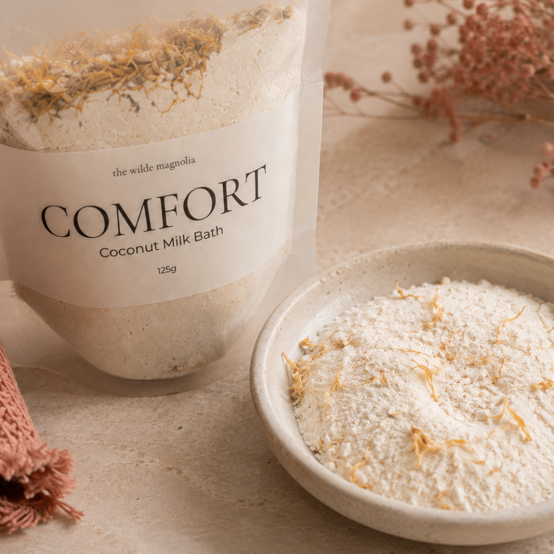 A clear stand-up pouch labeled 'COMFORT Coconut Milk Bath' with a white paper label, shown beside a small ceramic bowl filled with the white bath powder and dried botanical pieces. The scene is styled on a beige surface with a folded pink knit fabric and dried flowers in the background.