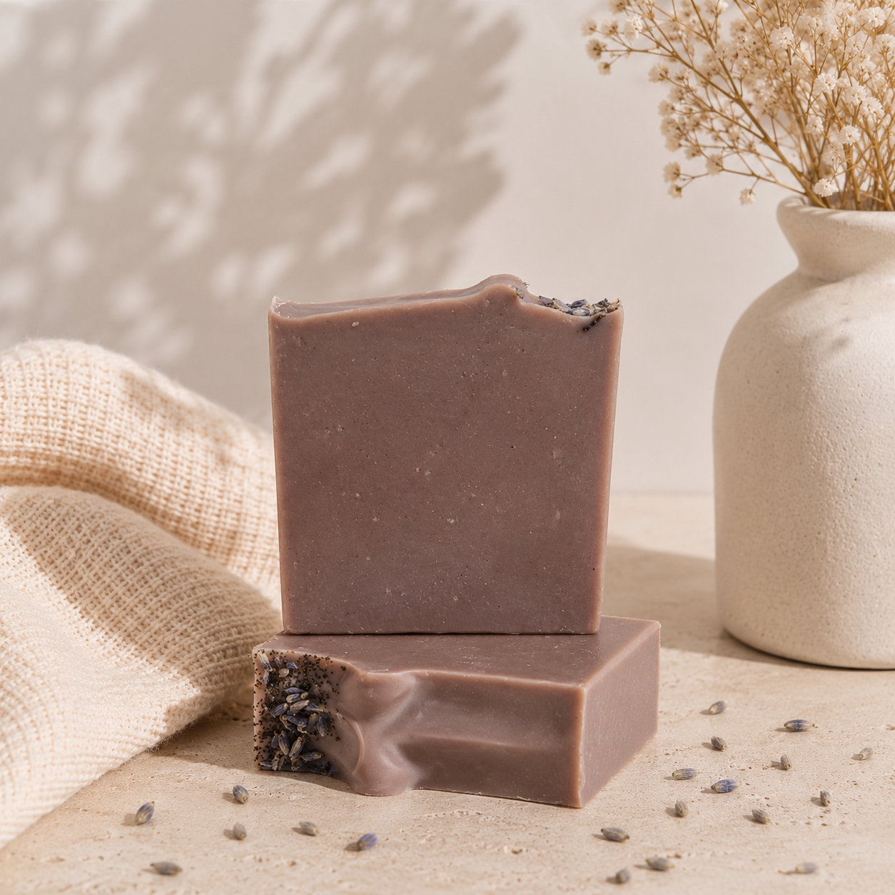 Two rectangular bars of handmade soap stacked on a beige surface, with one bar standing on the other to show the textured. The soap has a muted mauve-brown color and is topped with dried lavender.