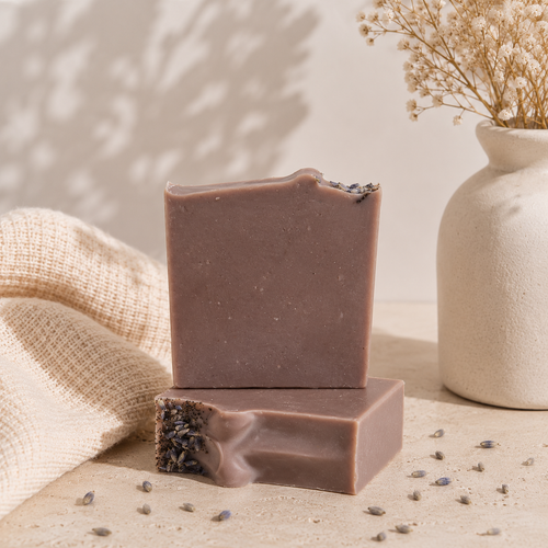 Two rectangular bars of handmade soap stacked on a beige surface, with one bar standing on the other to show the textured. The soap has a muted mauve-brown color and is topped with dried lavender.