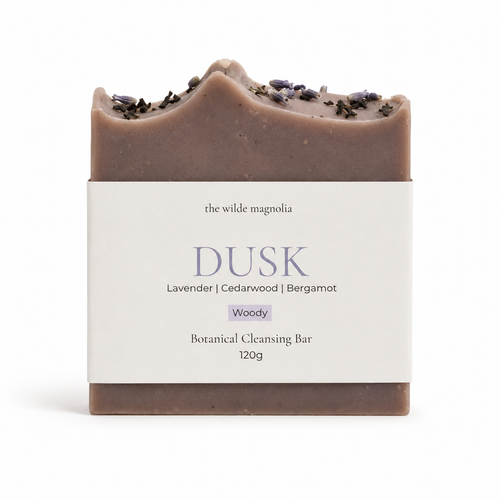 A single botanical cleansing bar wrapped in a white paper label, photographed on a clean white background. The bar is a muted dusty mauve-brown with dried lavender on top.