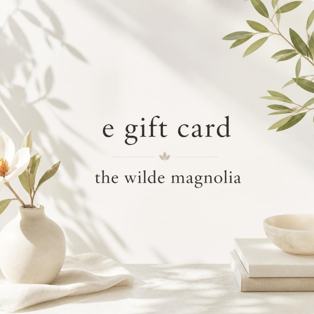 A gift card design with the text 'e gift card' and 'the wilde magnolia' centered on a warm off-white background with soft leaf shadows, plus decorative flowers, a vase, books, and a bowl.