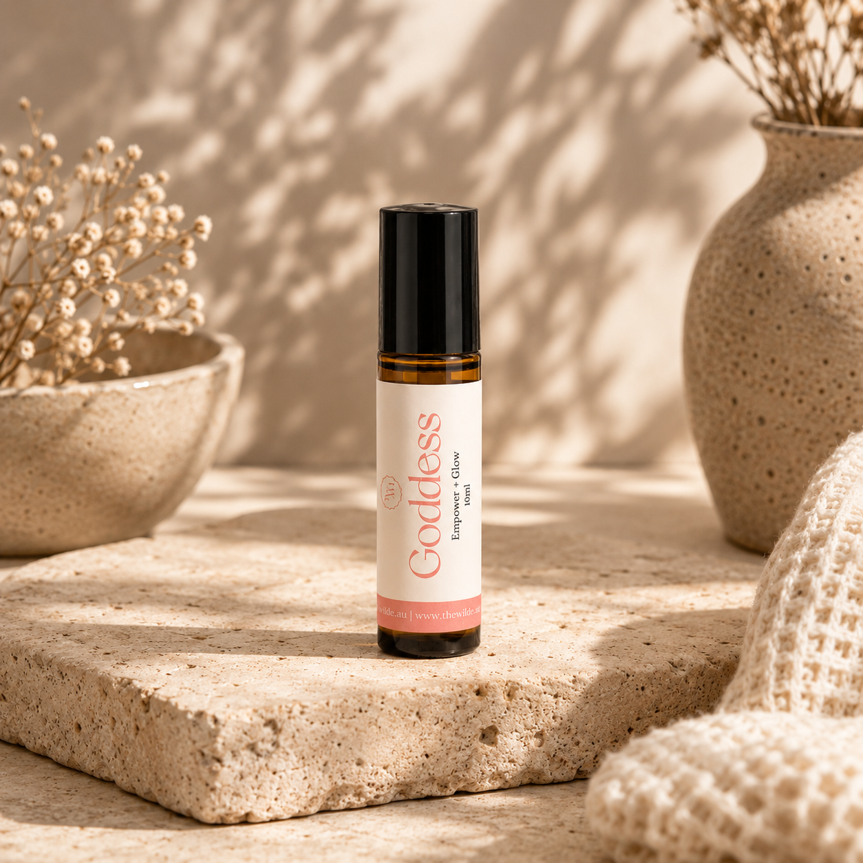 A small amber glass roller bottle labeled Goddess with a black cap, standing on a stone slab in a warm, sunlit setting with dried flowers and textured decor in the background.
