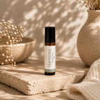 A small amber glass roller bottle with a black cap and a white label reading 'Harmony' stands on a textured stone slab in a warm, sunlit setting with dried flowers and ceramic vases in the background.