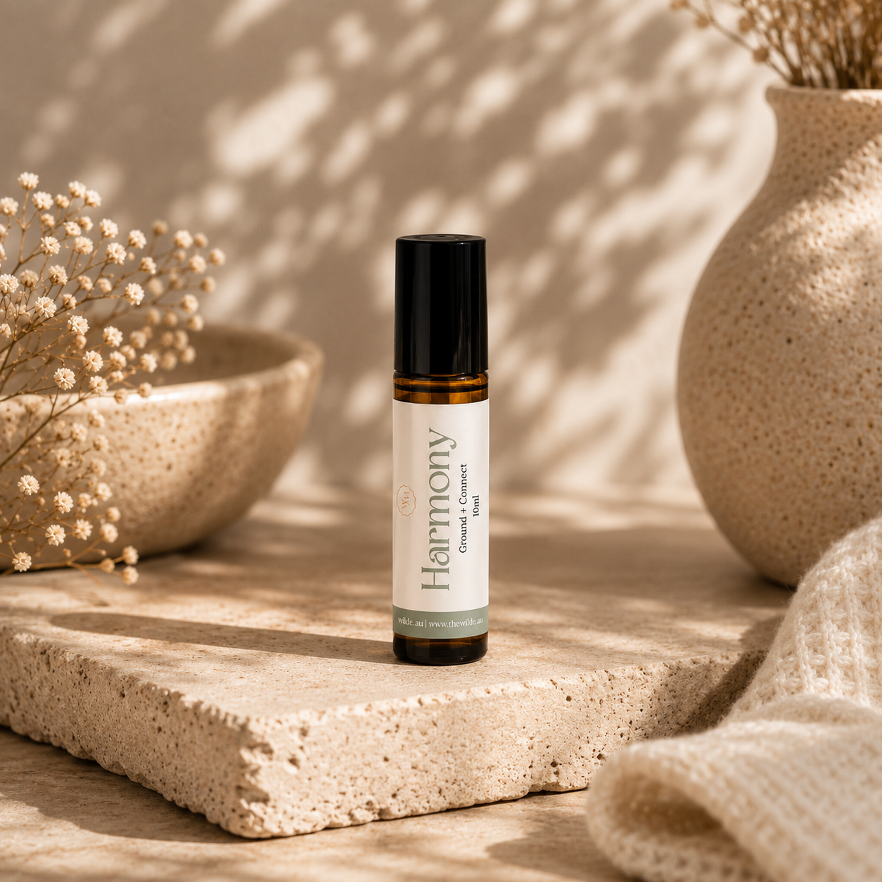 A small amber glass roller bottle with a black cap and a white label reading 'Harmony' stands on a textured stone slab in a warm, sunlit setting with dried flowers and ceramic vases in the background.