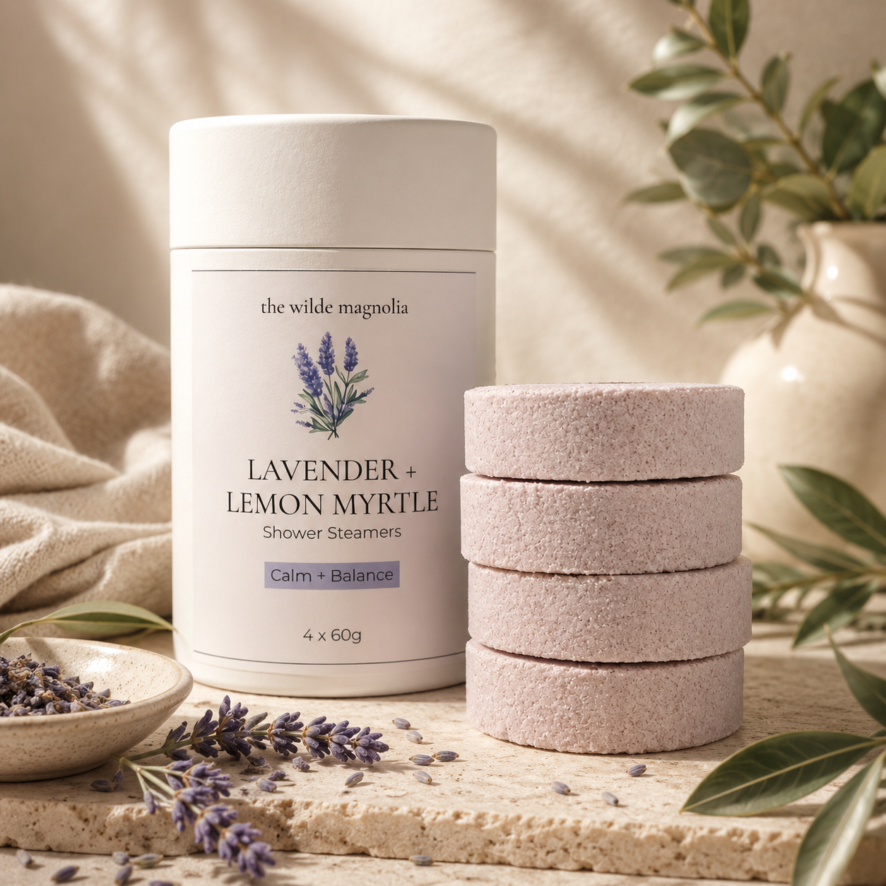 A white cylindrical tub of shower steamers labeled 'the wilde magnolia Lavender + Lemon Myrtle Shower Steamers' stands beside a stack of four pale purple steamers on a stone surface, with lavender flowers, green leaves, and soft neutral fabric in the background.