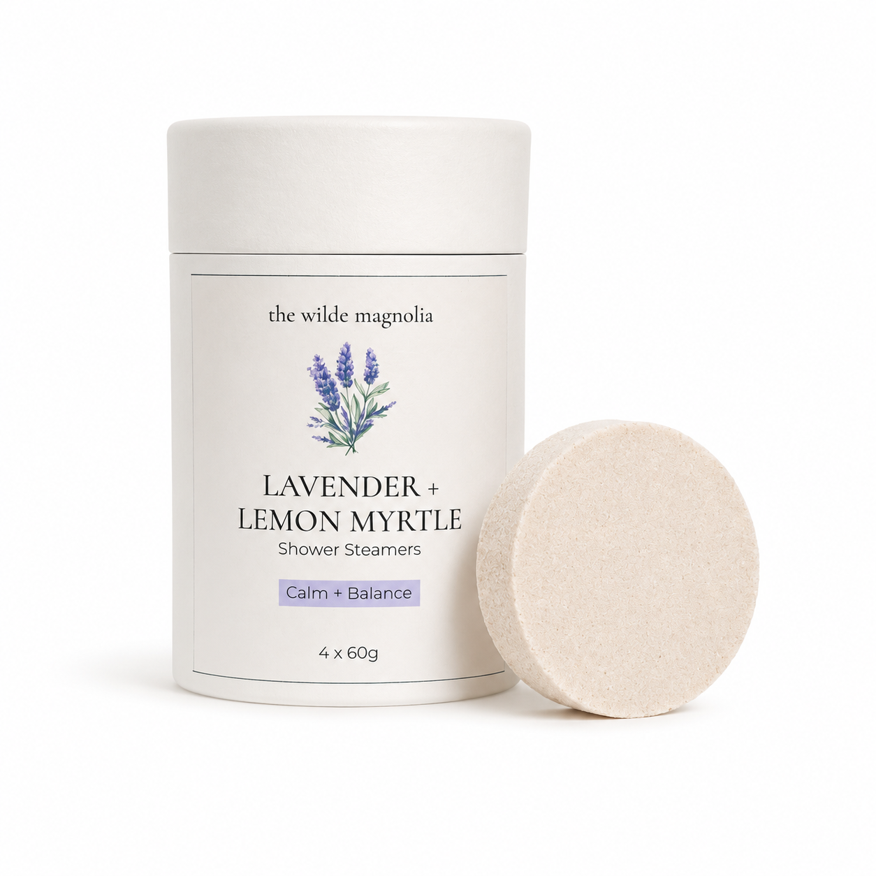 A white cylindrical container of 'the wilde magnolia' Lavender + Lemon Myrtle Shower Steamers, shown next to a single round pale purple steamer disc on a clean white background.