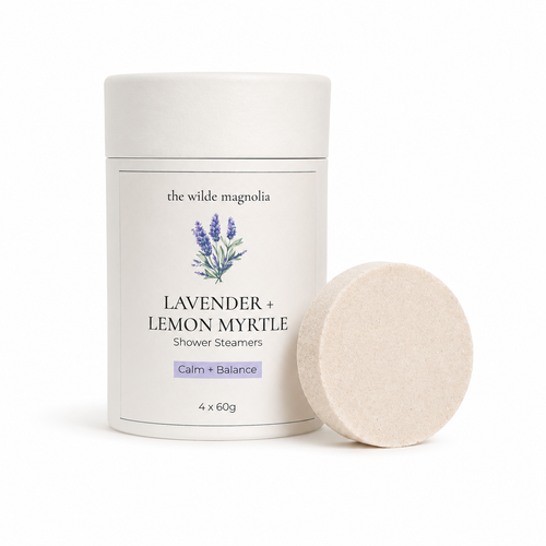 A white cylindrical container of 'the wilde magnolia' Lavender + Lemon Myrtle Shower Steamers, shown next to a single round pale purple steamer disc on a clean white background.