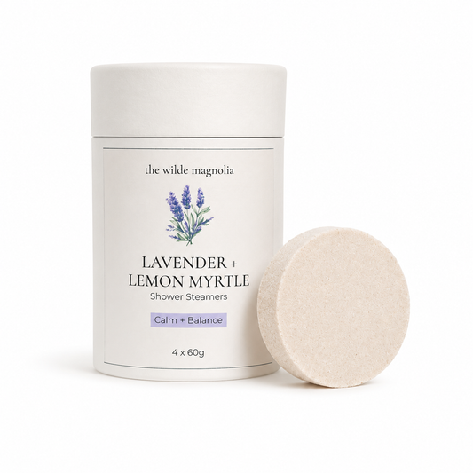 A white cylindrical container of 'the wilde magnolia' Lavender + Lemon Myrtle Shower Steamers, shown next to a single round pale purple steamer disc on a clean white background.