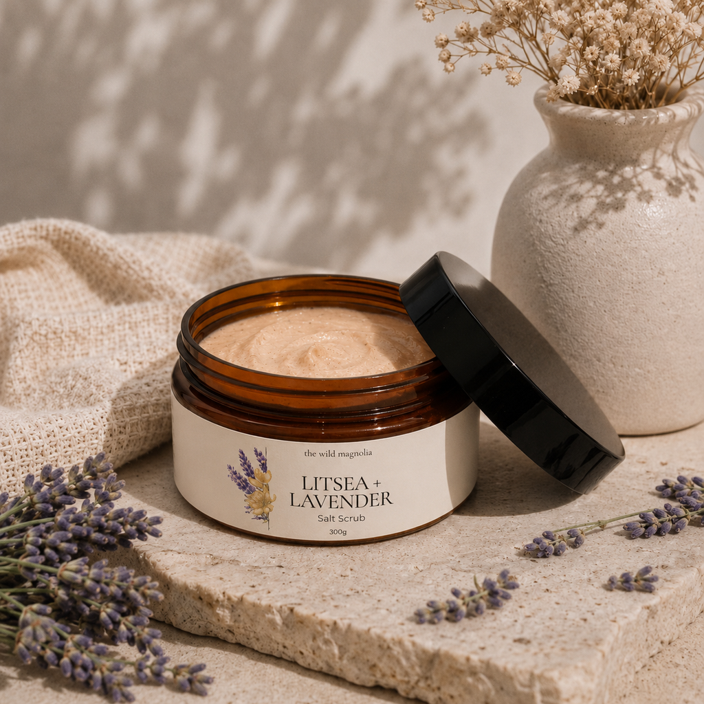 An open amber jar of Litsea + Lavender Salt Scrub sits on a stone slab with its black lid leaning beside it. The cream-colored scrub is visible inside, with dried lavender sprigs, a beige fabric cloth, and a textured ceramic vase in the background under soft shadow lighting.