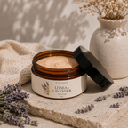 An open amber jar of Litsea + Lavender Salt Scrub sits on a stone slab with its black lid leaning beside it. The cream-colored scrub is visible inside, with dried lavender sprigs, a beige fabric cloth, and a textured ceramic vase in the background under soft shadow lighting.