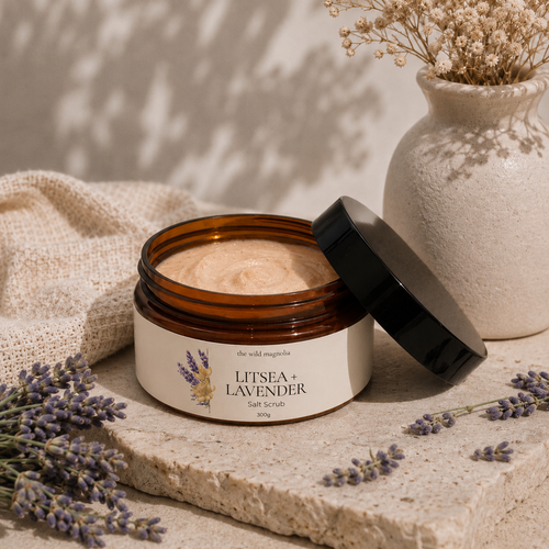 An open amber jar of Litsea + Lavender Salt Scrub sits on a stone slab with its black lid leaning beside it. The cream-colored scrub is visible inside, with dried lavender sprigs, a beige fabric cloth, and a textured ceramic vase in the background under soft shadow lighting.