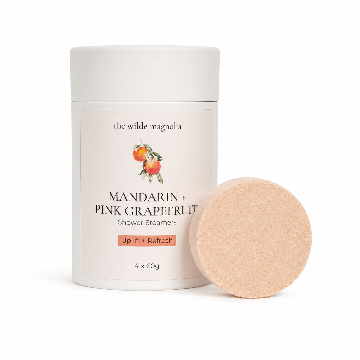 A white cylindrical container of shower steamers with a pale orange shower steamer puck leaning against it. The label reads 'the wilde magnolia' and 'MANDARIN + PINK GRAPEFRUIT Shower Steamers,' with a small fruit illustration.