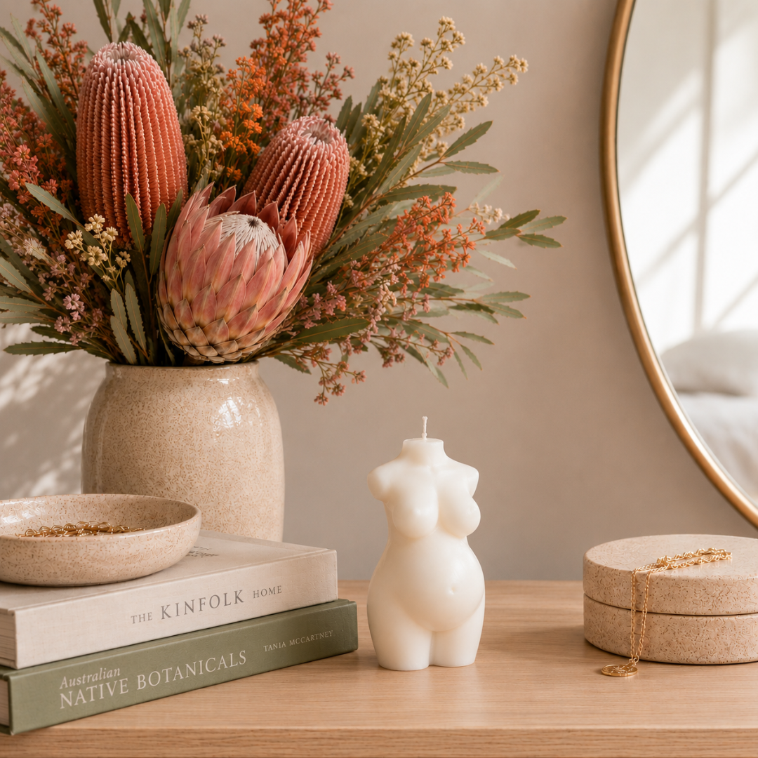 A styled wooden dressing table with a ceramic vase of dried Australian native flowers, stacked books, and a white sculptural candle shaped like a pregnant torso, a small bowl, and a round jewelry box with a gold chain.