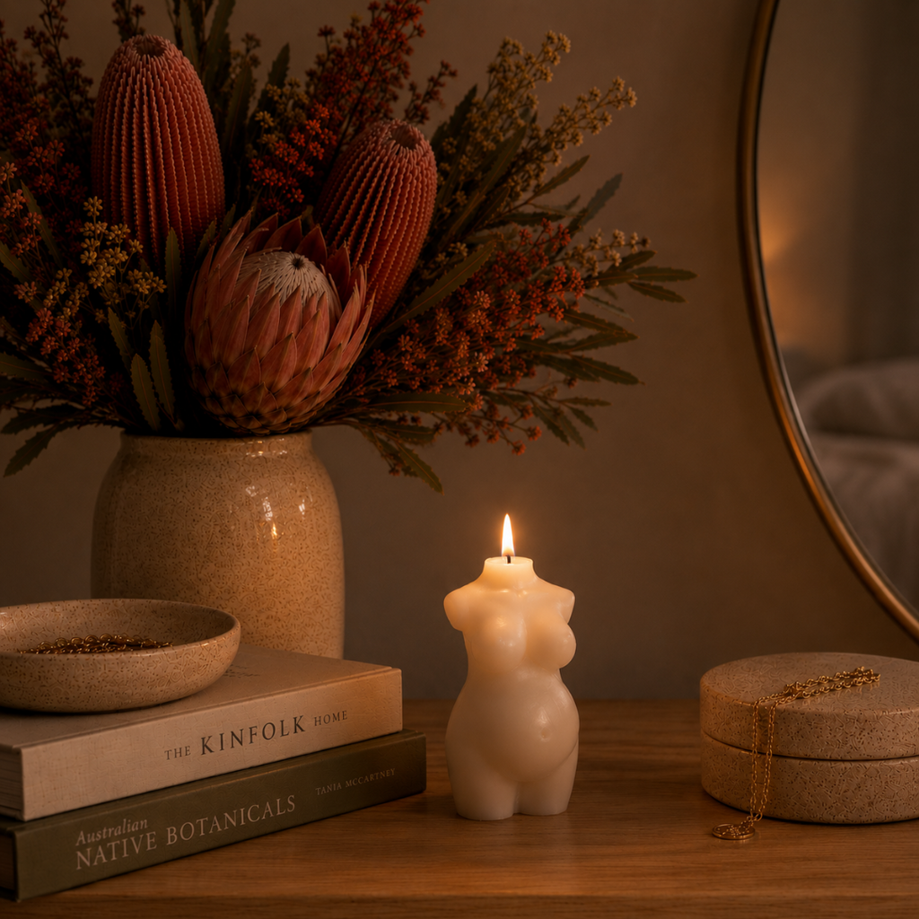 A lit cream-colored candle shaped like a pregnant torso sits on a wooden table next to a stack of books, a vase of dried protea flowers, and a round mirror in a warm, cozy room setting.