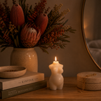 A lit cream-colored candle shaped like a pregnant torso sits on a wooden table next to a stack of books, a vase of dried protea flowers, and a round mirror in a warm, cozy room setting.