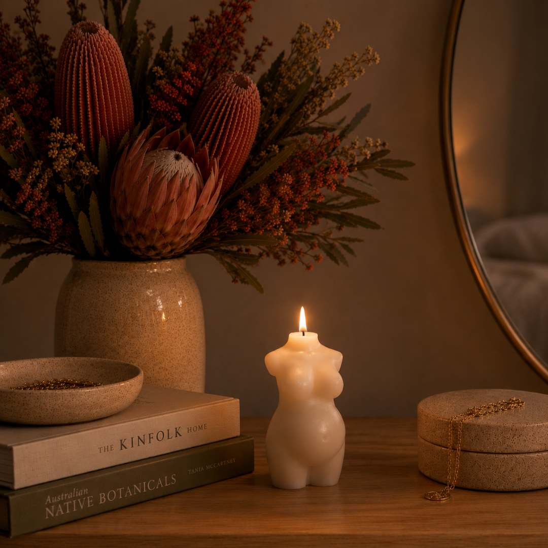A lit cream-colored candle shaped like a pregnant torso sits on a wooden table next to a stack of books, a vase of dried protea flowers, and a round mirror in a warm, cozy room setting.