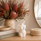 A styled tabletop scene with a white female-form bust candle centered in front of a ceramic vase filled with dried protea flowers and rust-toned filler stems. A round mirror, stacked books, a small bowl, and a round jewelry box with a gold chain are also visible on a light wood surface.