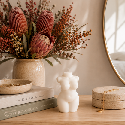 A styled tabletop scene with a white female-form bust candle centered in front of a ceramic vase filled with dried protea flowers and rust-toned filler stems. A round mirror, stacked books, a small bowl, and a round jewelry box with a gold chain are also visible on a light wood surface.