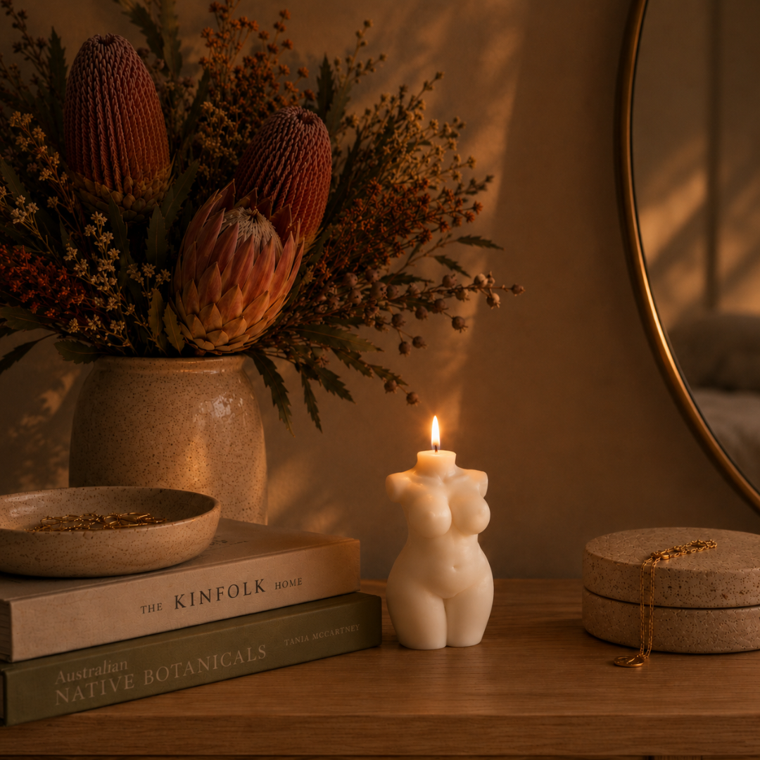 A lit white candle shaped like a curvy female torso sits on a wooden table beside a vase of protea flowers, a round mirror, and a small jewelry box in a warm, softly lit interior setting.