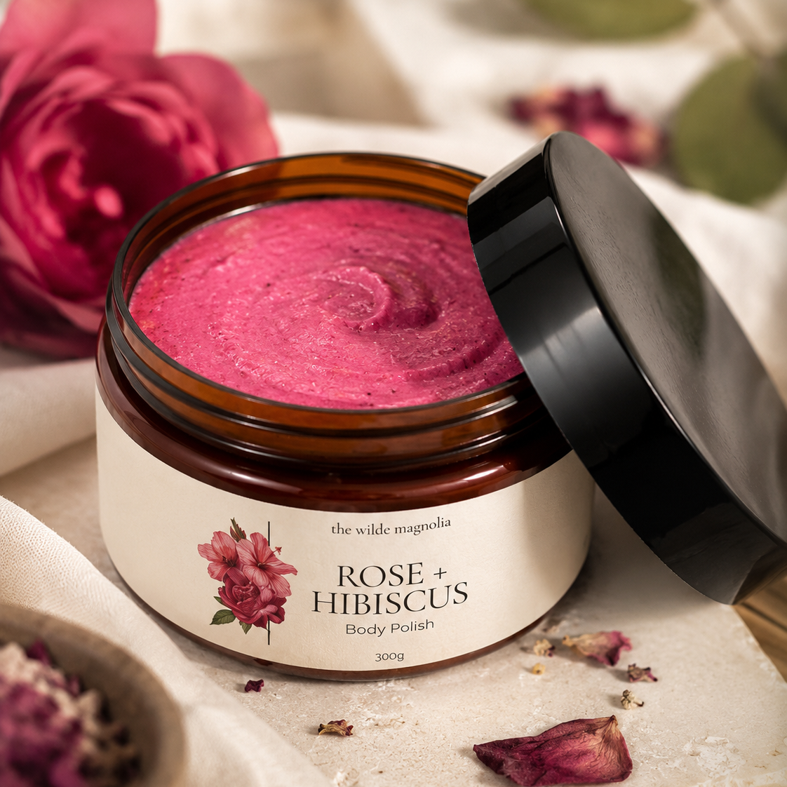 An open amber glass jar of body polish with a black lid resting beside it on a soft fabric surface. The product inside is a vivid pink scrub, and the label reads 'the wilde magnolia ROSE + HIBISCUS Body Polish 300g'.