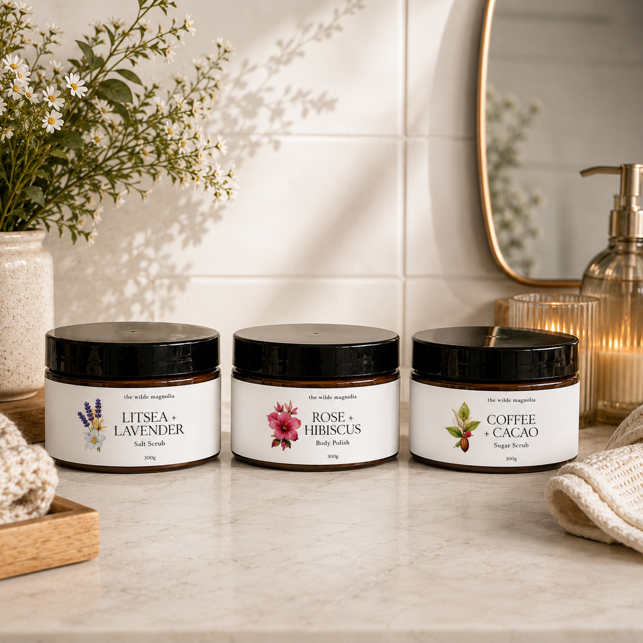 Three jars of 'The Wile Magnolia's' body scrubs arranged on a marble countertop in a bathroom setting. The jars are labeled 'Litsea + Lavender', 'Rose + Hibiscus', and 'Coffee + Cacao' with black lids and white labels featuring botanical illustrations.