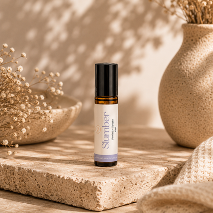 A 10ml amber glass roller bottle with a black cap and a cream label reading 'Slumber' stands on a textured stone slab. The scene is styled with dried flowers, a woven textile, and a speckled ceramic vase in warm natural light with leafy shadows.
