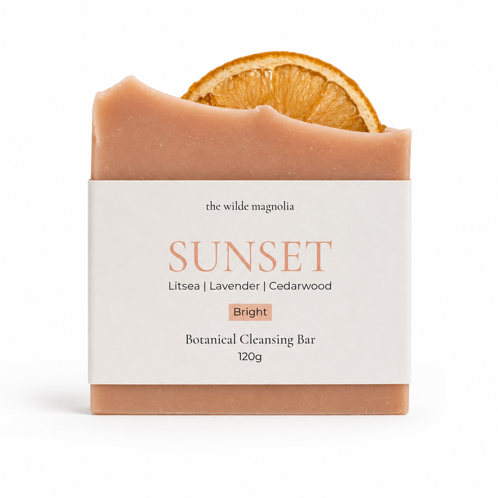A botanical cleansing bar from The Wilde Magnolia, named 'Sunset', featuring a dusty peach-colored bar with a dried orange slice embedded at the top, wrapped in a white paper label.