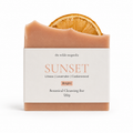 A botanical cleansing bar from The Wilde Magnolia, named 'Sunset', featuring a dusty peach-colored bar with a dried orange slice embedded at the top, wrapped in a white paper label.