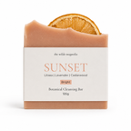 A botanical cleansing bar from The Wilde Magnolia, named 'Sunset', featuring a dusty peach-colored bar with a dried orange slice embedded at the top, wrapped in a white paper label.