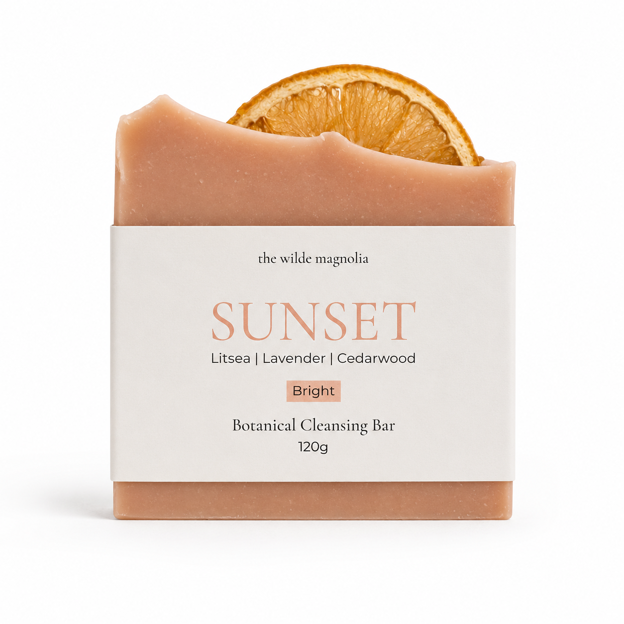 A botanical cleansing bar from The Wilde Magnolia, named 'Sunset', featuring a dusty peach-colored bar with a dried orange slice embedded at the top, wrapped in a white paper label.