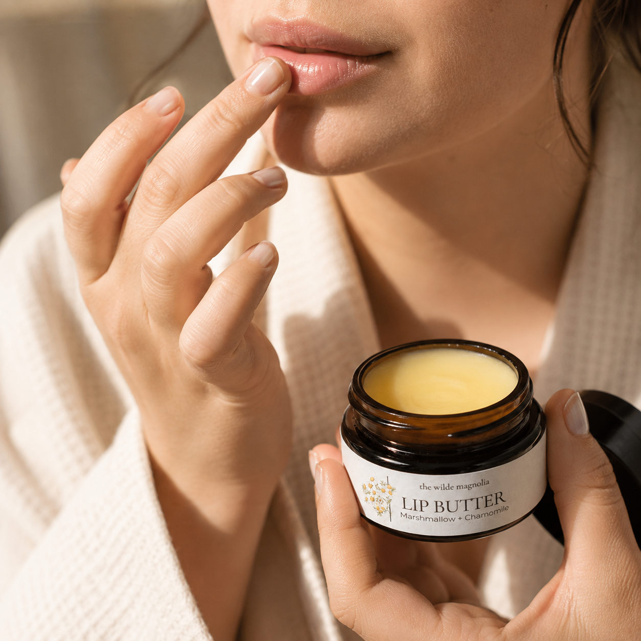 A close-up lifestyle shot of a woman applying lip balm from an amber glass jar. She is wearing a white textured robe, and the jar is labeled 'the wild magnolia' Lip Butter with Marshmallow and Chamomile.