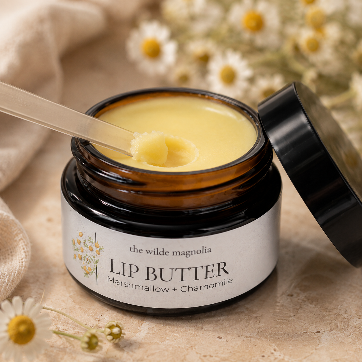 An open amber glass jar of lip balm with a black lid leaning against it, shown on a beige surface with chamomile flowers. The label reads 'the wilde magnolia LIP BUTTER Marshmallow + Chamomile.